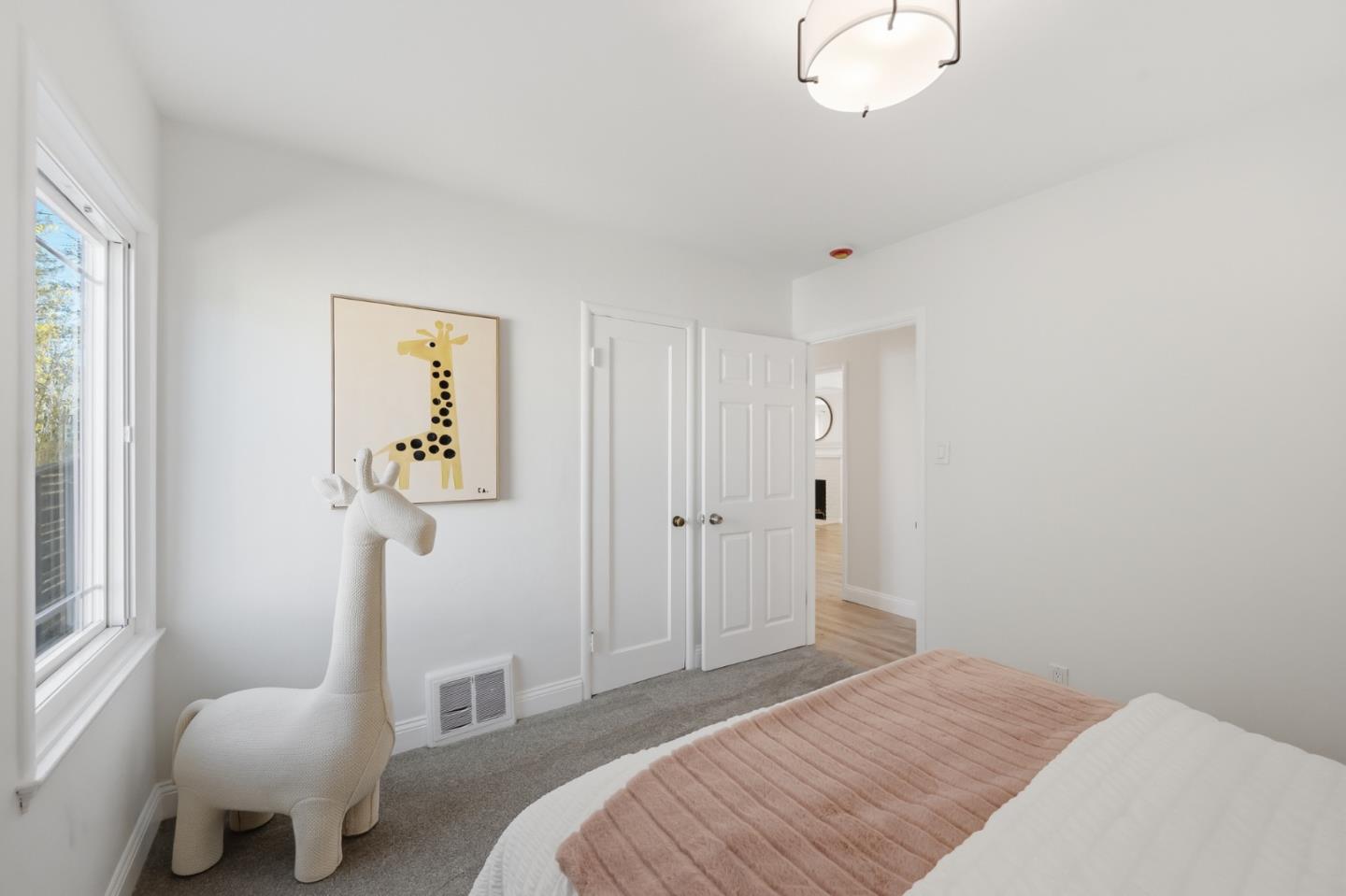 Bedroom with a pink quilt, large window, and white giraffe-shaped chair beneath giraffe artwork.
