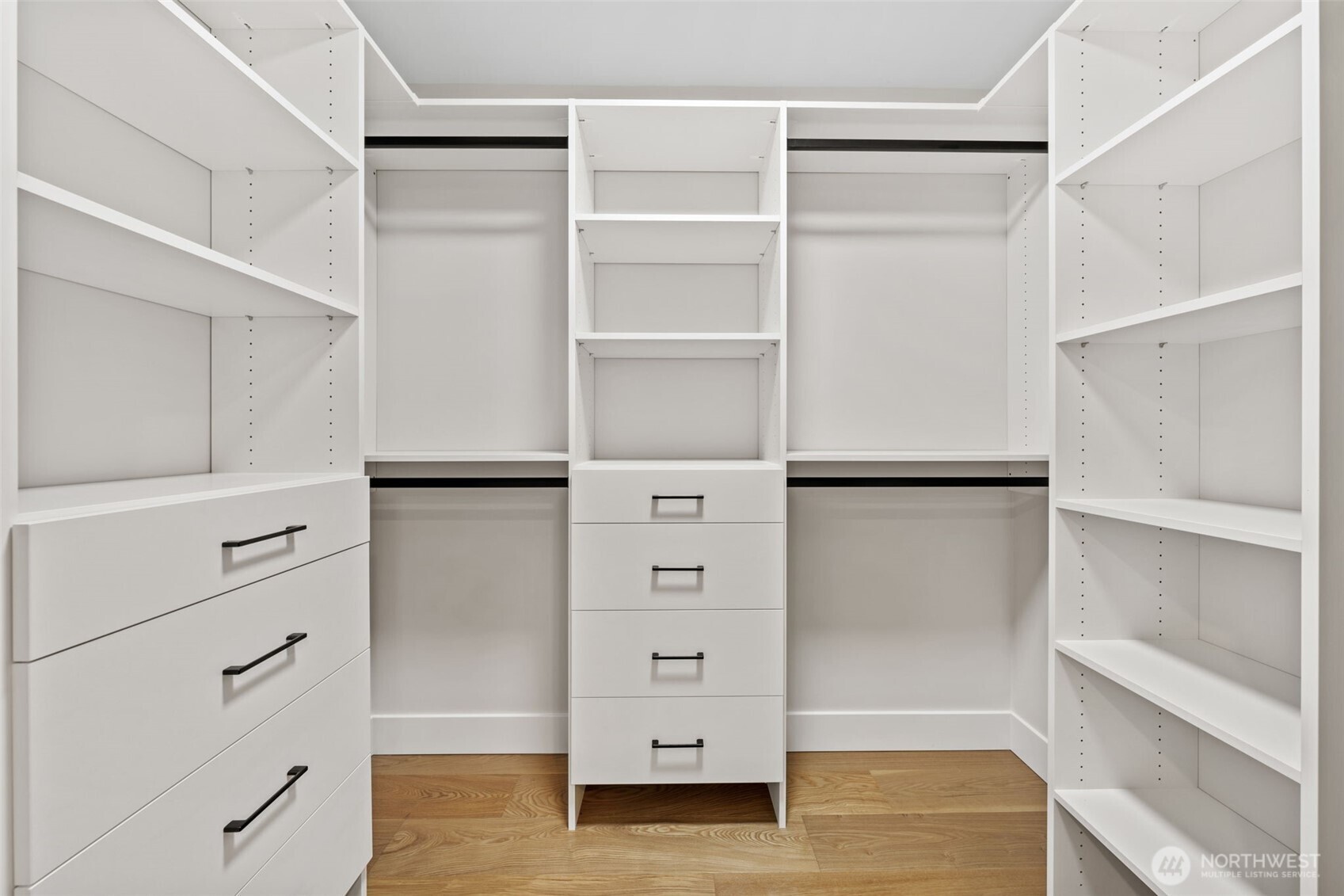 Walk-in closet with white built-in shelving and drawers, black pulls, and wood flooring.