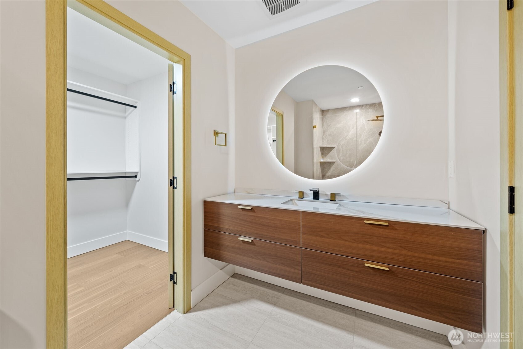 Bathroom vanity with a round illuminated mirror beside an open walk-in closet.