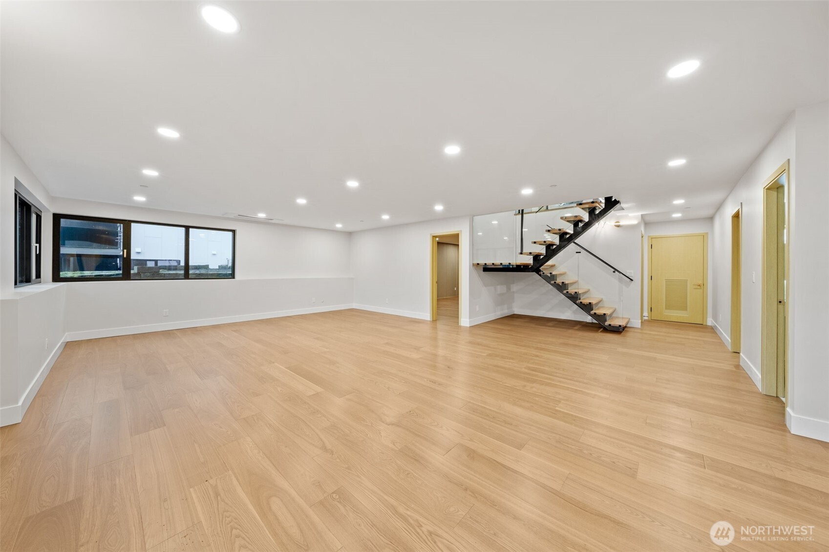Empty living room with light wood floors, large windows, and a modern floating staircase.
