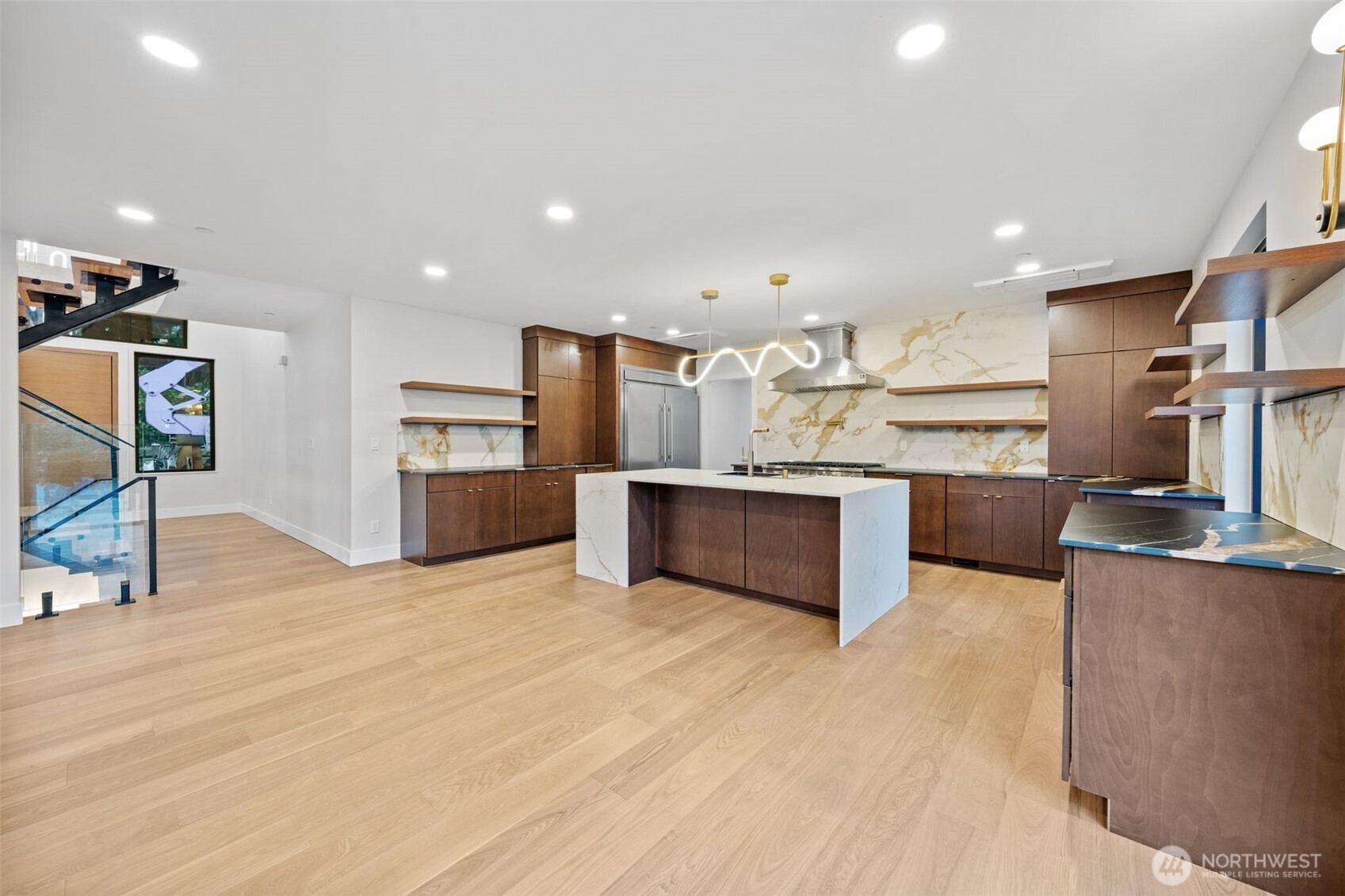 Modern kitchen with a large marble island, walnut cabinetry, and a glass-railed staircase.
