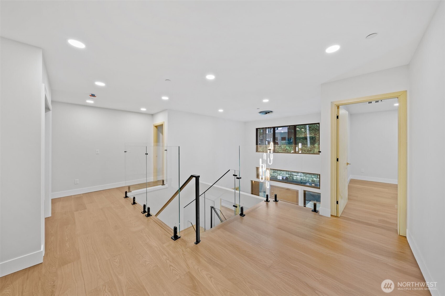 Upper-floor landing with light wood flooring, glass stair railings, and a tall window.