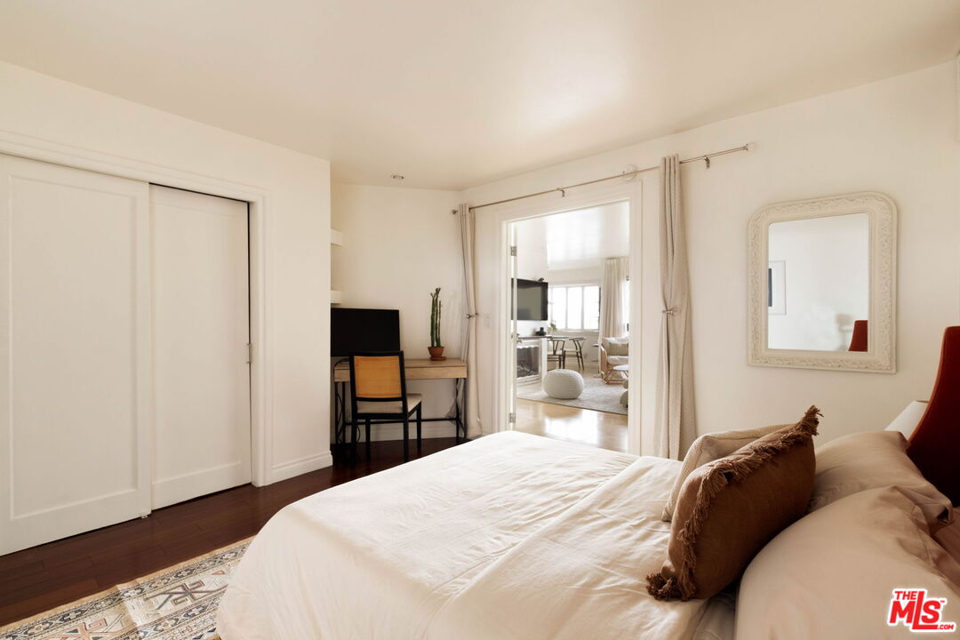 Bedroom with a white bed, patterned rug, desk, and doorway to a bright living area.