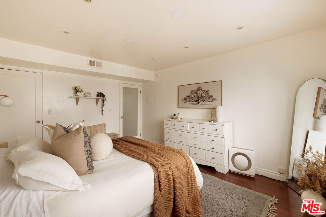 Staged bedroom with a white bed, rust-colored knit throw, white dresser, and arched mirror.