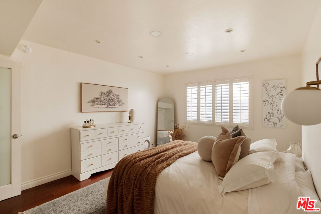 Furnished bedroom with a white bed, brown knitted throw, and white dresser beneath plantation shutters.