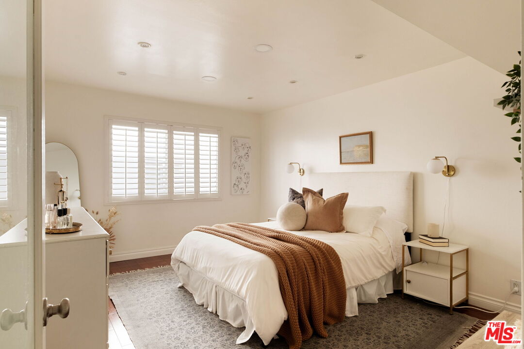 Staged bedroom with a white bed, rust-colored throw, and plantation shutters.