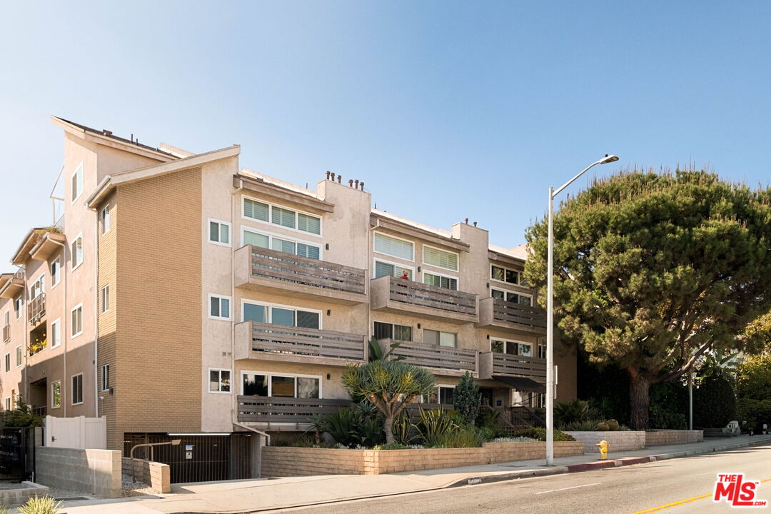 Beige apartment building with stacked balconies, landscaped frontage, and a gated garage entrance.