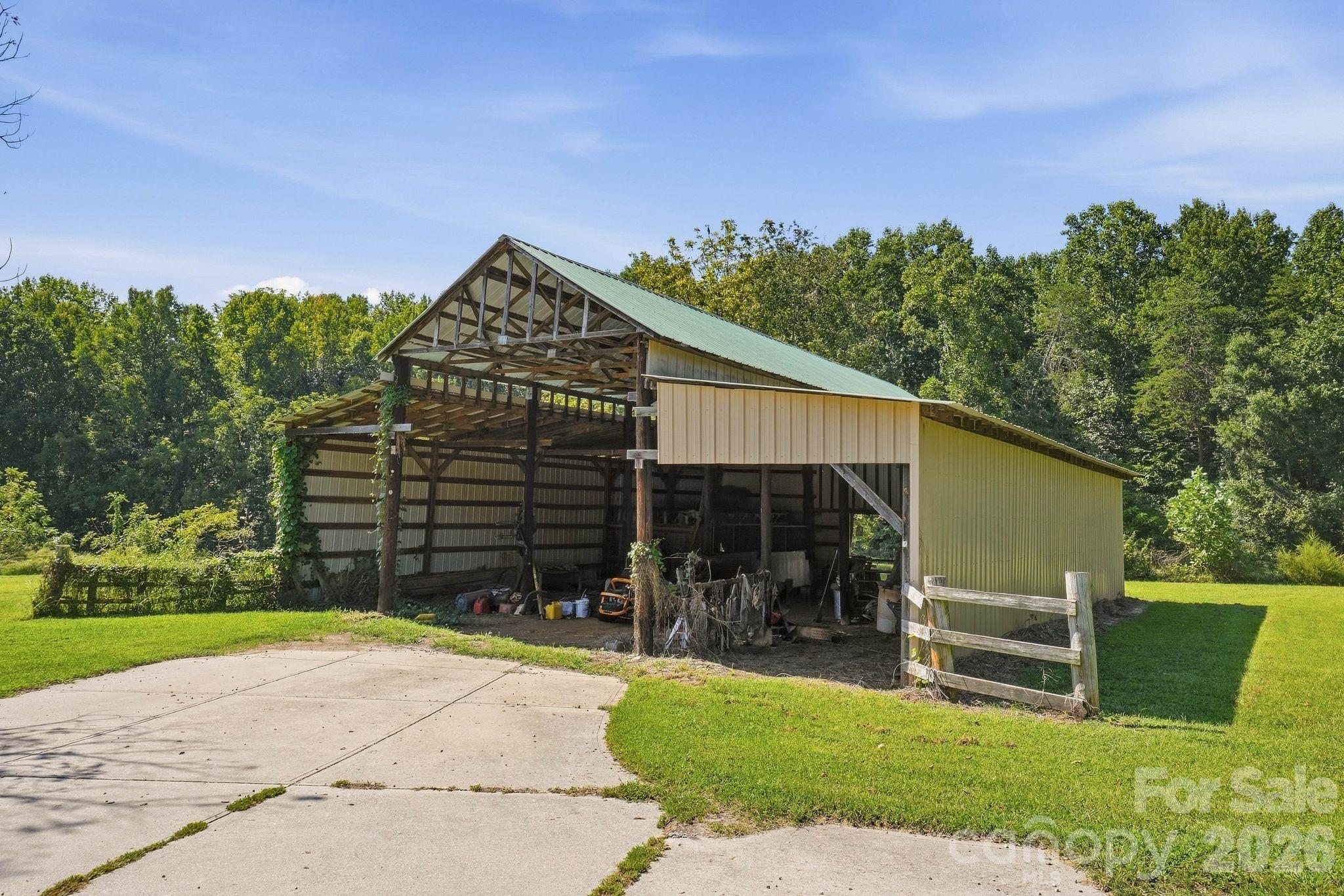 Open-sided wooden barn with a green metal roof beside a concrete driveway.