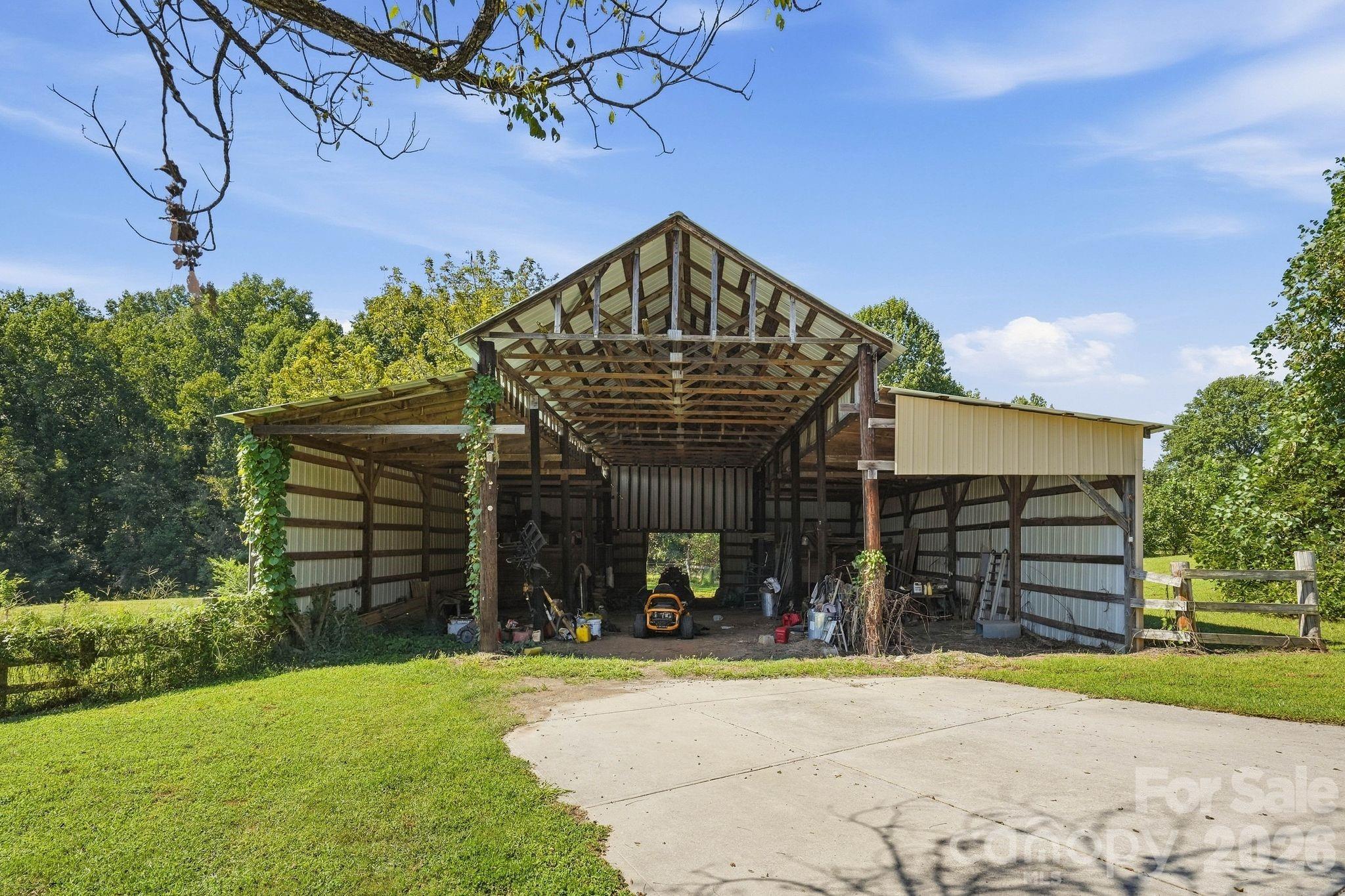 Open-sided timber-frame barn with corrugated metal panels beside a concrete driveway and grassy yard.