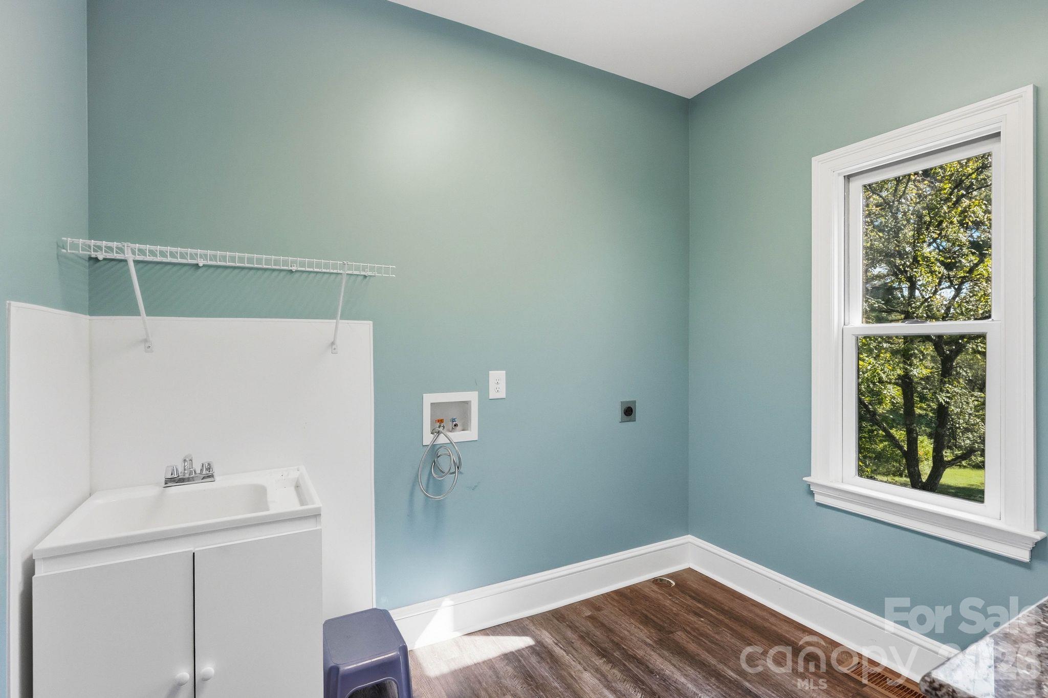 Empty laundry room with a utility sink, washer hookups, drying rack, and window overlooking trees.