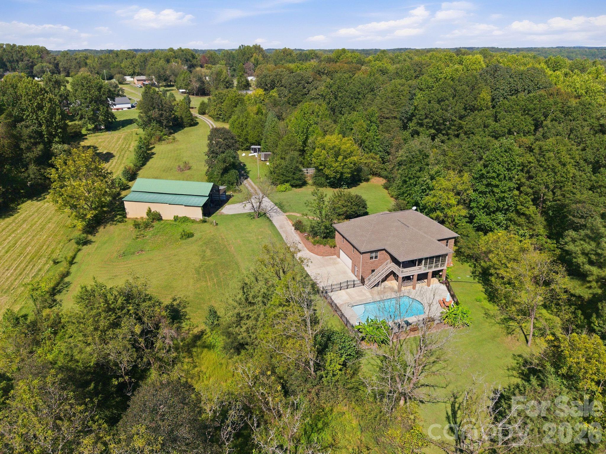 Aerial view of a brick home and swimming pool surrounded by lawns and trees, with a detached outbuilding nearby.