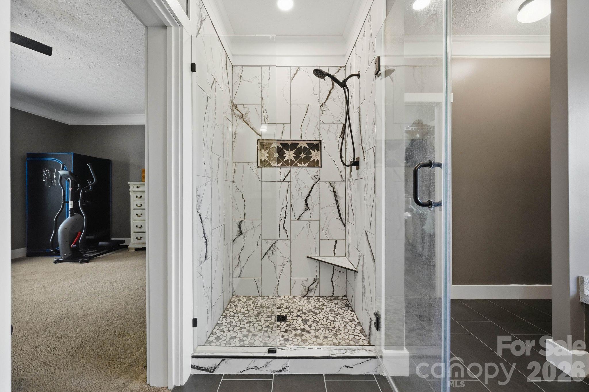 Walk-in shower with veined tile, black fixtures, and a pebble-stone floor beside an exercise room.