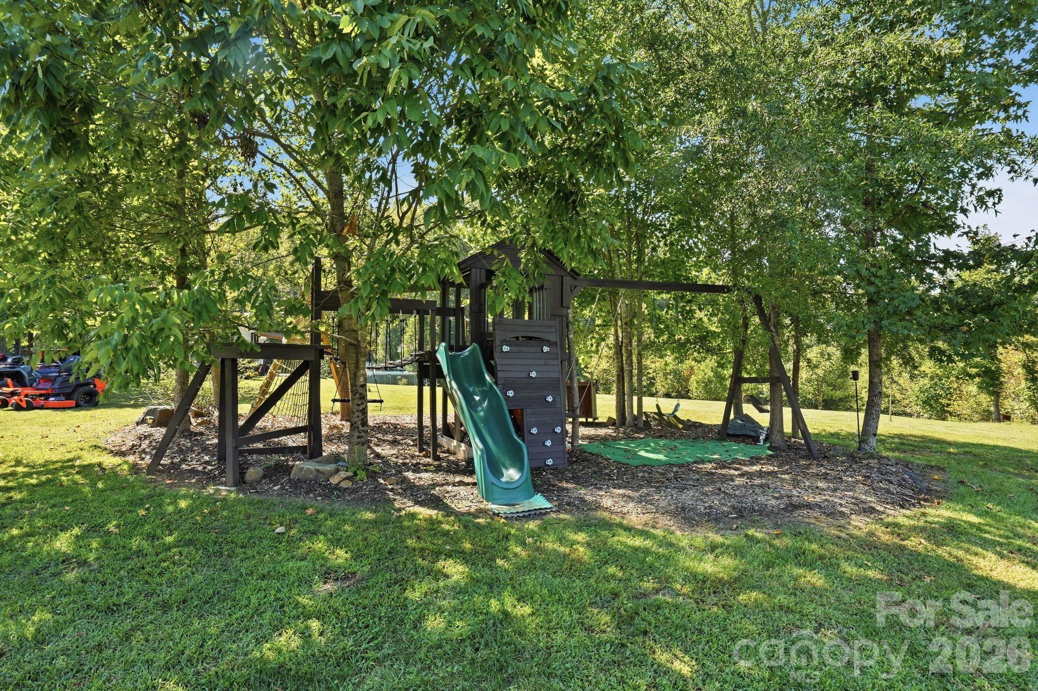 Backyard playset with a green slide, climbing wall, and swings beneath mature trees.