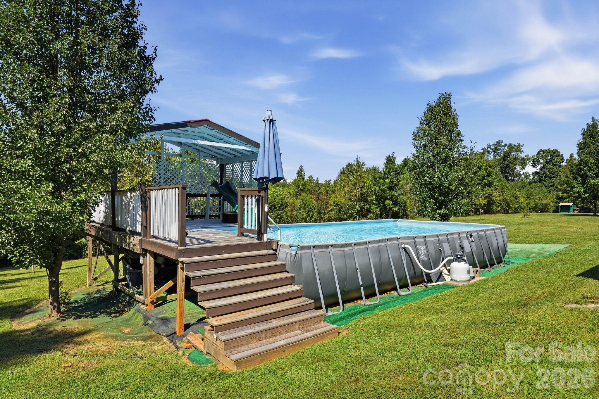 Above-ground pool beside a raised wooden deck with stairs and a slide in a tree-lined lawn.