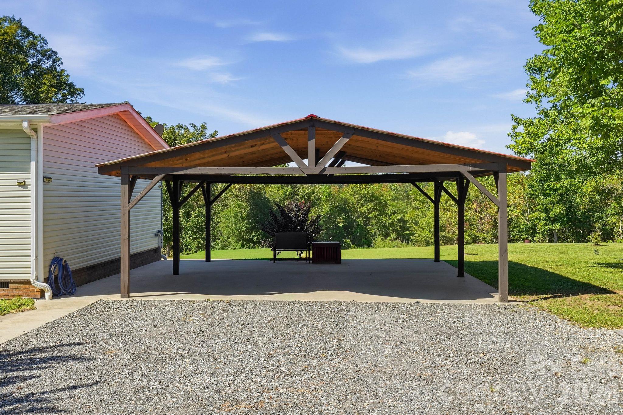 Open-sided wooden carport beside a light-colored house, with a gravel driveway and lawn.