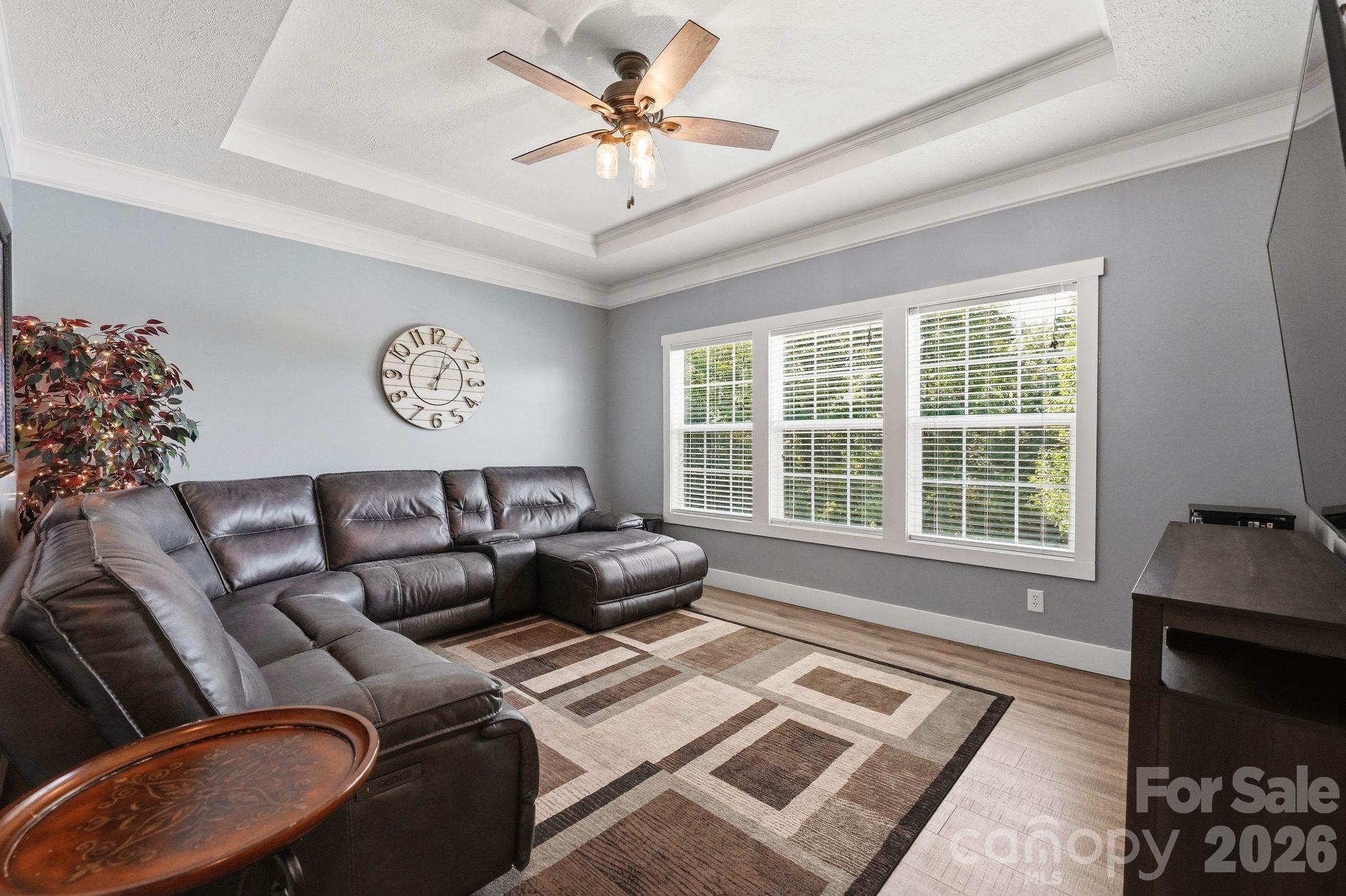 Living room with a dark leather sectional, geometric rug, tray ceiling, and three bright windows.