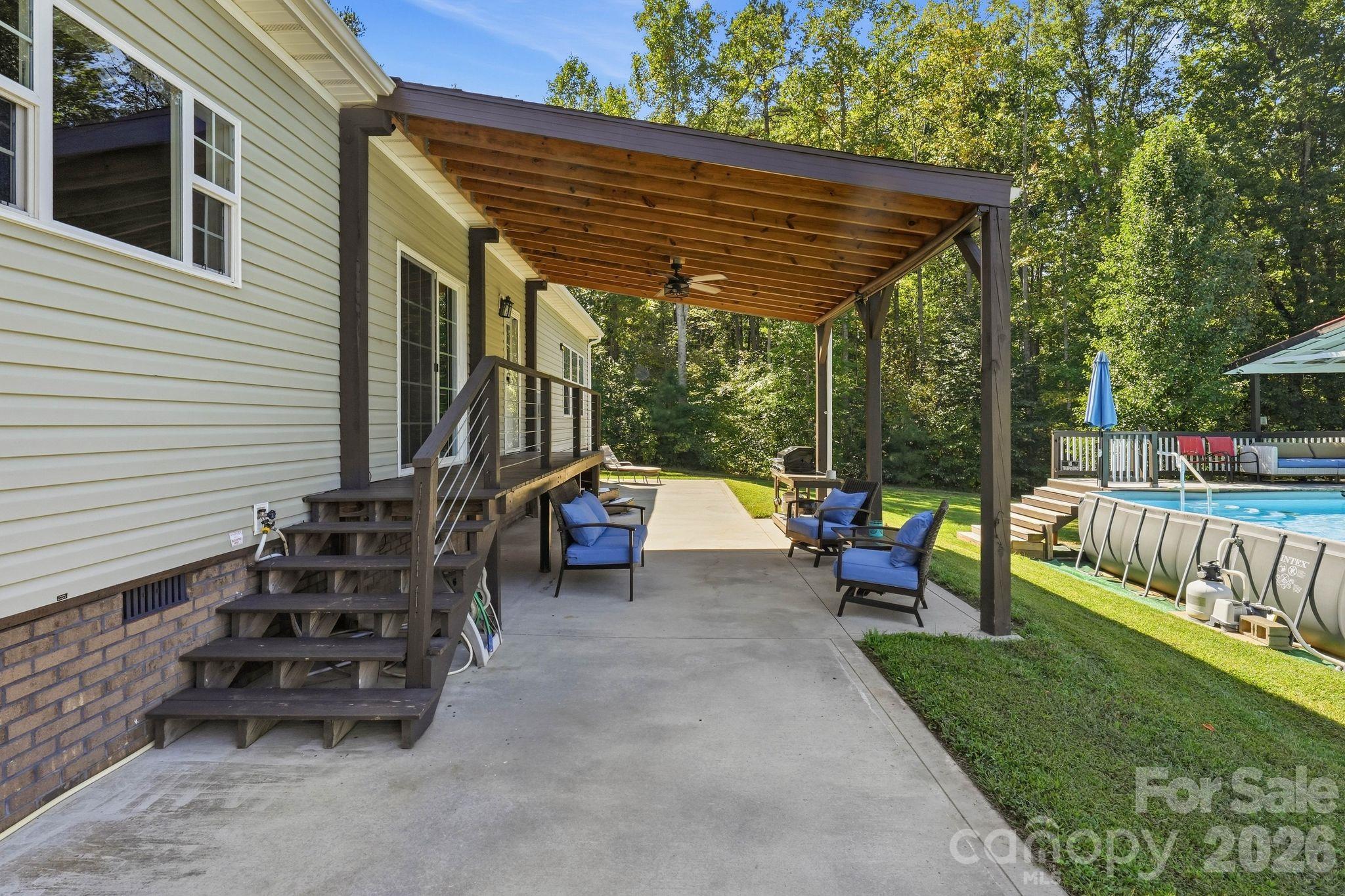 Covered patio with wooden pergola, blue lounge chairs, and an above-ground pool beside the house.