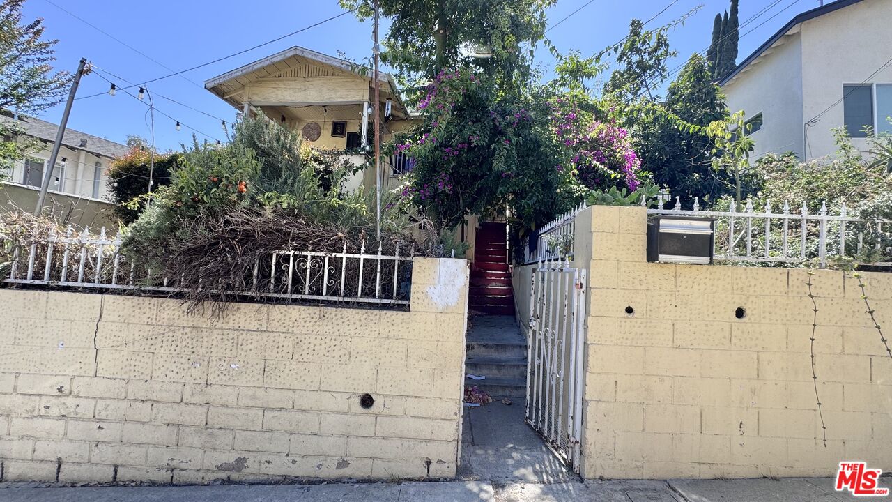 Street view of 1065 N Gage Ave, a two-story home behind a cream-colored wall and metal gate, with steps leading to the entrance beneath flowering bougainvillea.