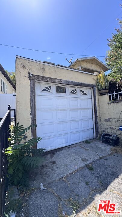 White garage door and cracked driveway at 1065 N Gage Ave, with a small stucco garage and plants along the side.