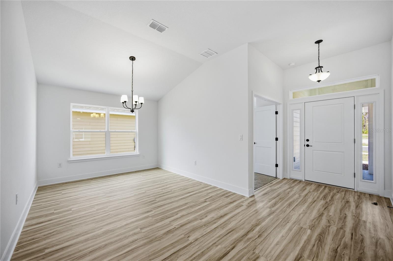 A bright, empty dining room featuring light wood-look flooring, a vaulted ceiling, and a chandelier, visible from the front entryway.