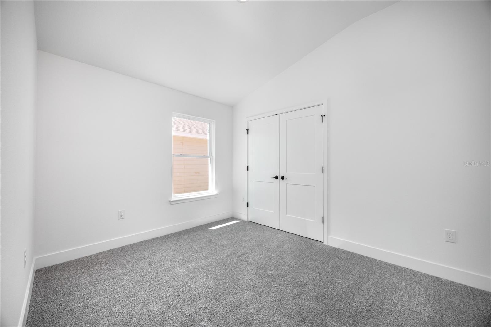 An empty bedroom with white walls, grey carpeting, a single window, and closed double-door closet.