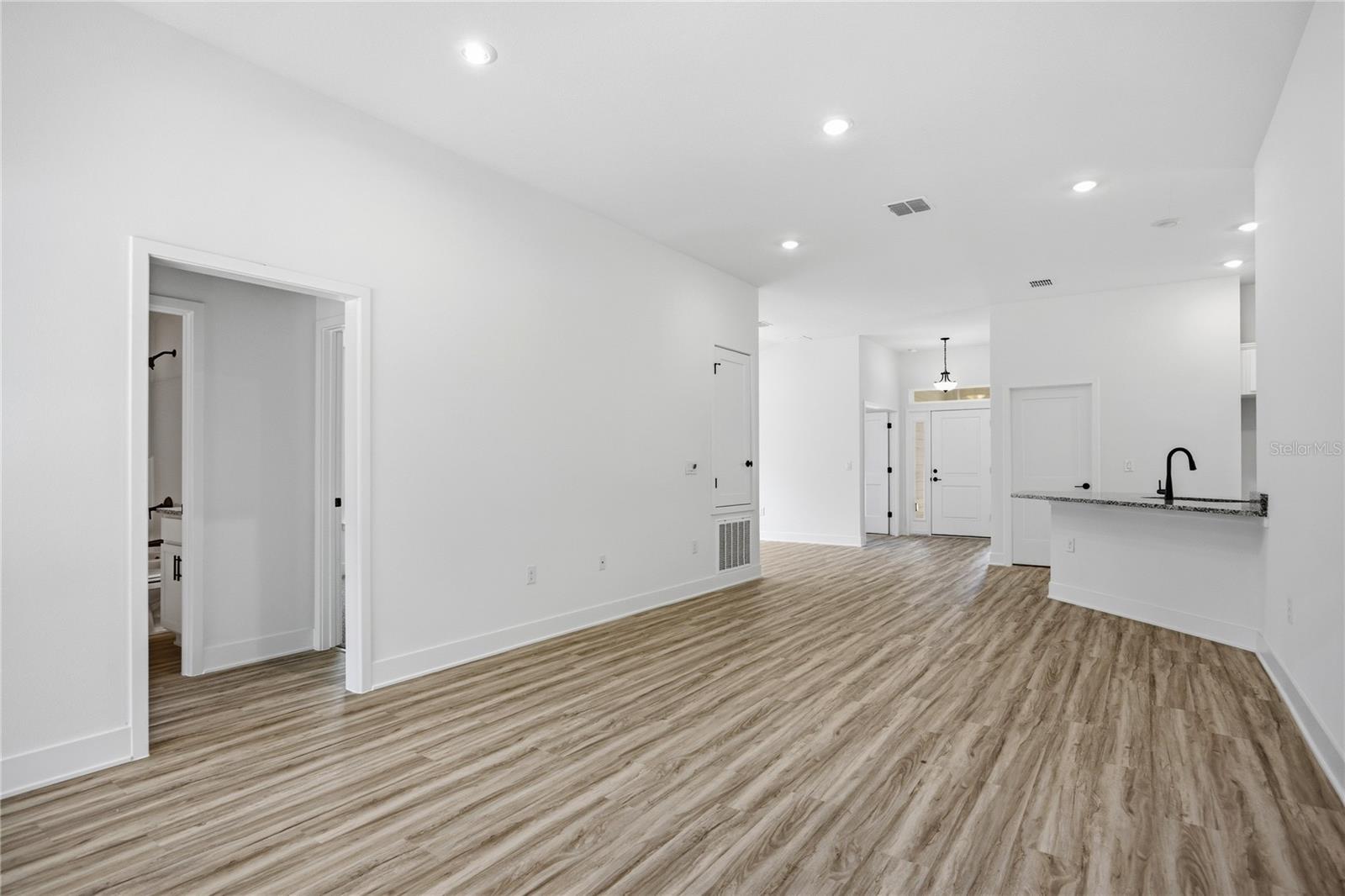 A wide-angle view of a modern living room space featuring light wood-look floors, white walls, and a glimpse into the kitchen area with a breakfast bar.