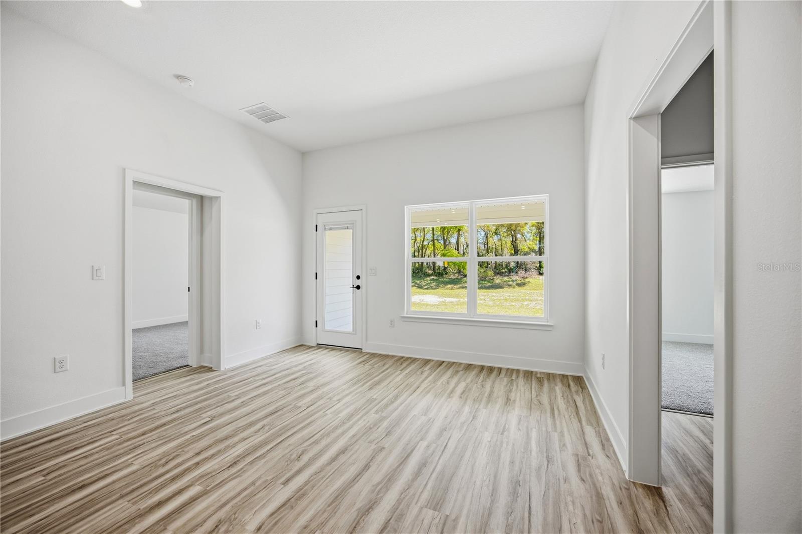 A vacant, brightly lit living room with wood-look flooring, white walls, and two doorways leading to carpeted rooms.