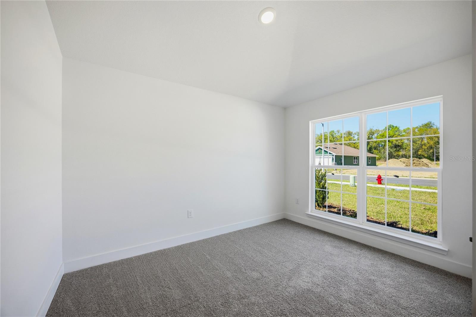 A vacant bedroom with neutral walls, grey carpeting, and a large window looking out onto a residential street.