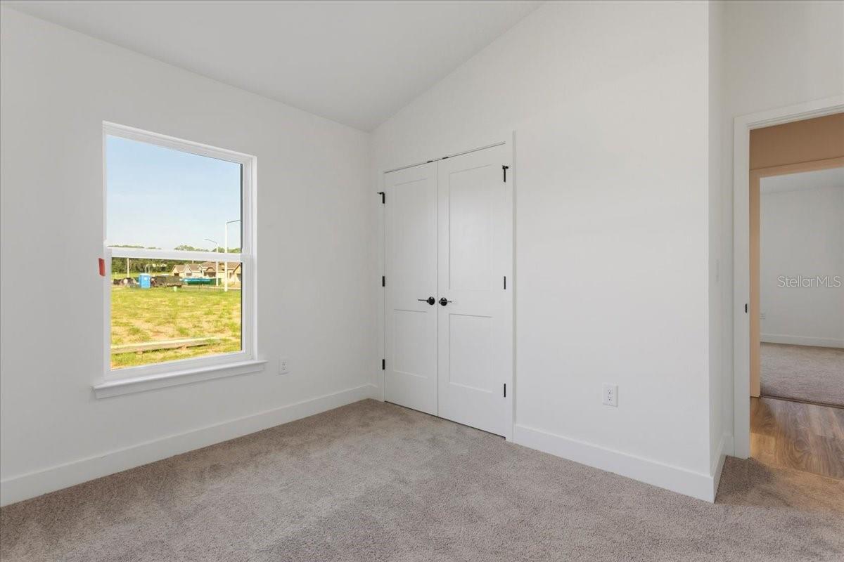 A vacant bedroom with white walls, gray carpet, a closet with white doors, and a window looking out onto a grassy area.