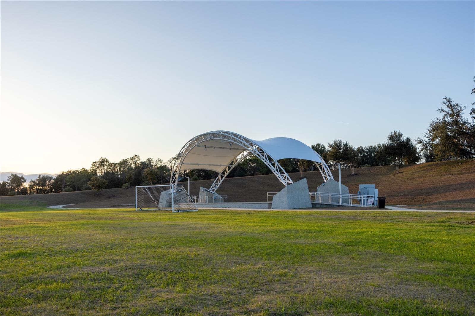 An outdoor amphitheater stage structure with a white tension fabric roof sits on a grassy field in a park setting.