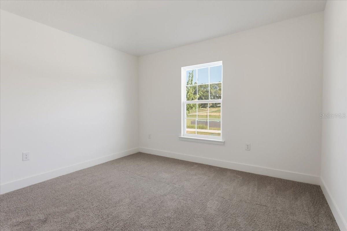 An empty bedroom with white walls, gray carpet, and a single window showing a grassy outdoor view.