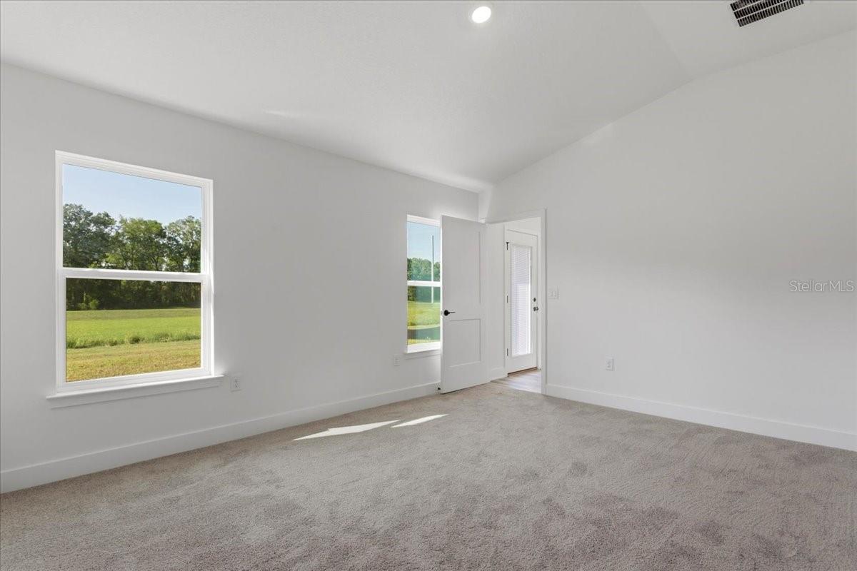 An empty bedroom with white walls, tan carpet, and windows looking out onto a grassy field.