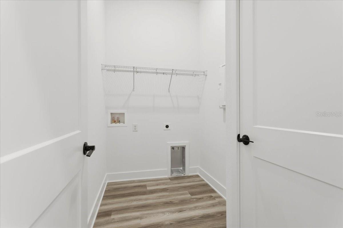 A small white laundry closet with wire shelving and laundry hookups, featuring wood-look vinyl flooring.