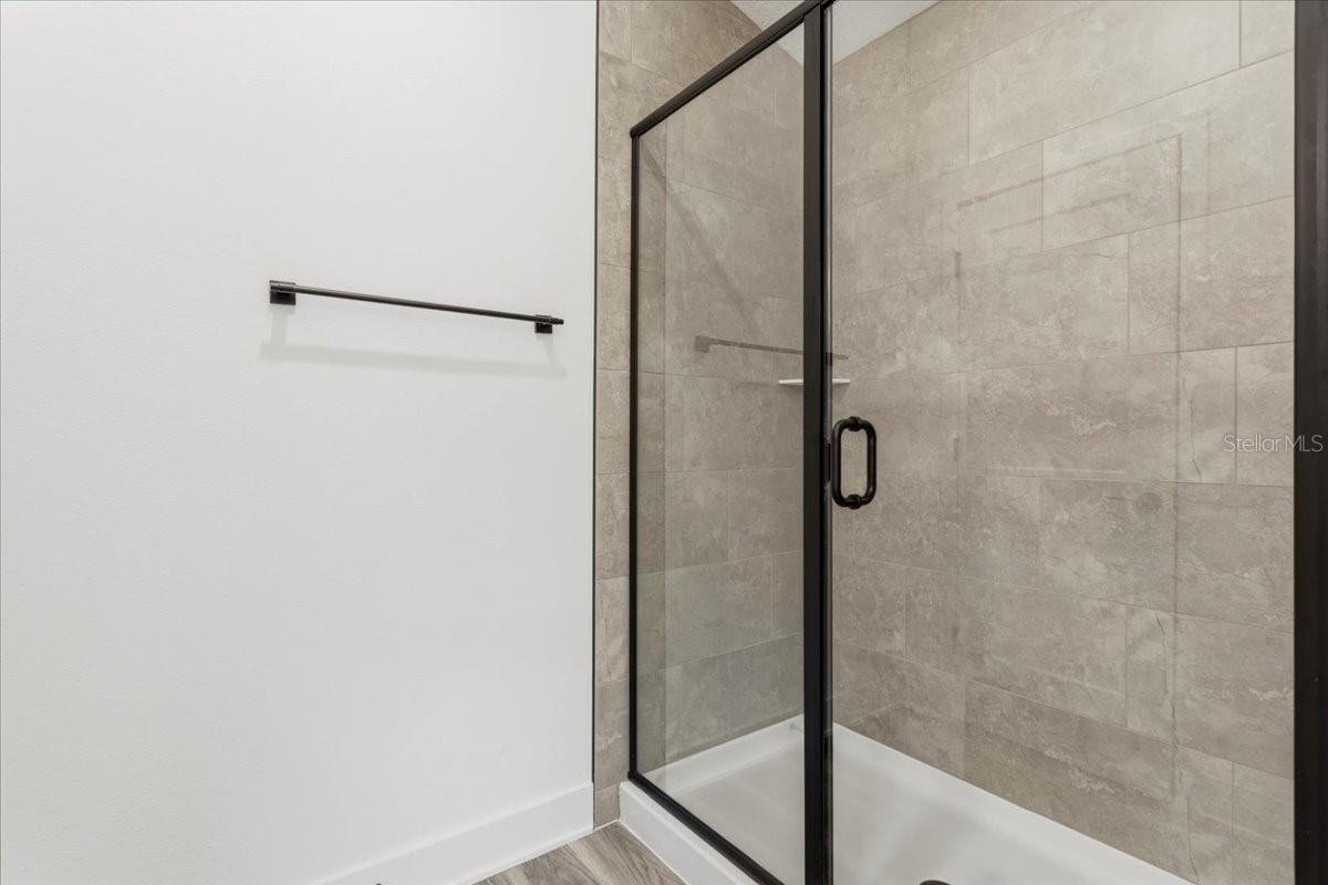 A modern walk-in shower with grey tiled walls and black metal frame glass doors, next to a white wall with a black towel bar.