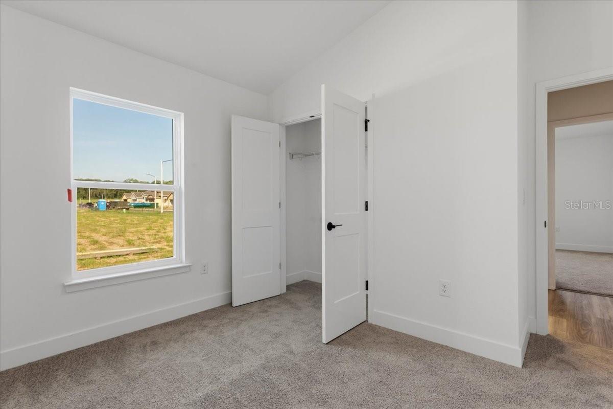 A vacant bedroom with gray carpet, white walls, and an open closet door looking out towards an empty lot.