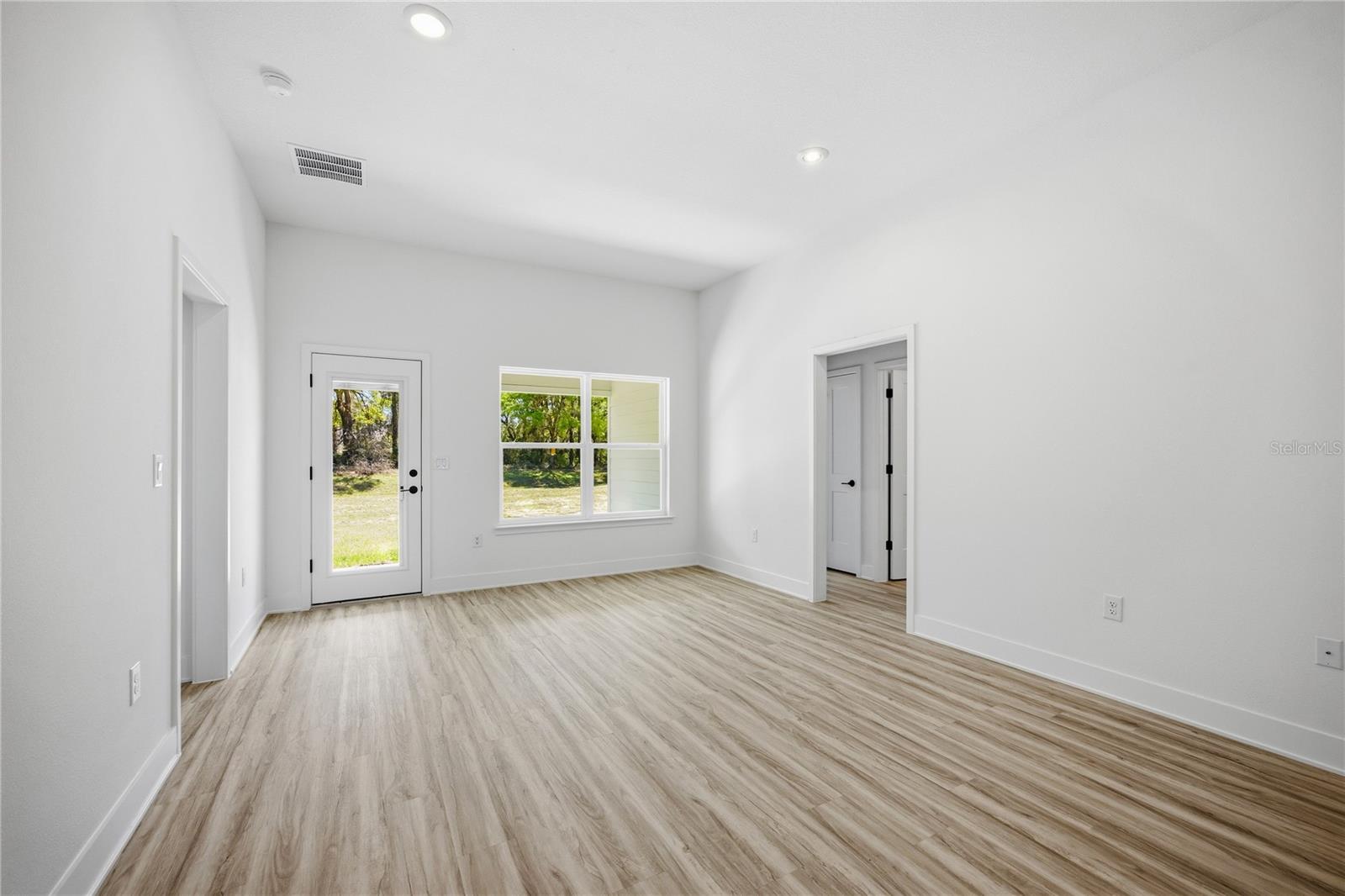 A vacant living room featuring white walls, light wood-look vinyl plank flooring, a glass-paneled exterior door, and large windows looking out to trees.