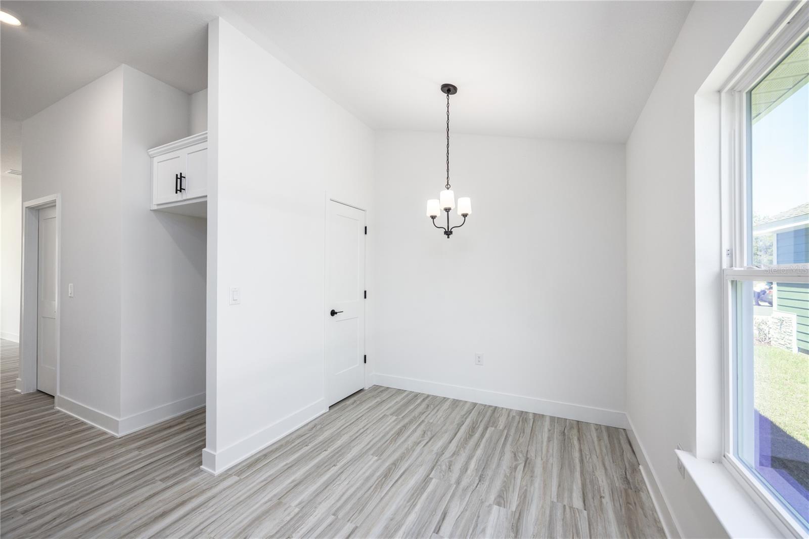 A clean, bright, empty dining area featuring grey-toned wood-look plank flooring, white walls, a modern chandelier, and a large window.