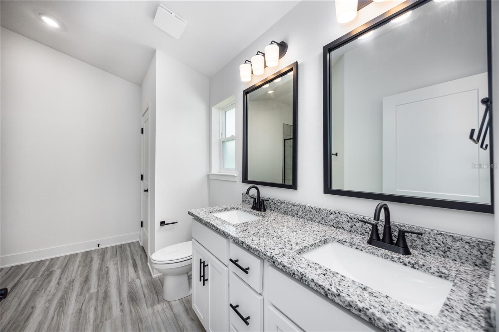 A clean modern bathroom featuring a white vanity with granite countertop, dual black-framed mirrors, and wood-look laminate flooring.