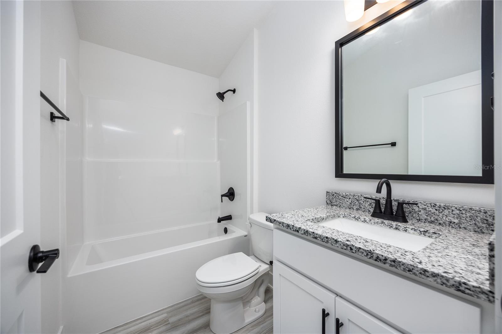 Modern bathroom featuring white walls, a granite-topped vanity, a black-framed mirror, and a clean white bathtub/shower combination.