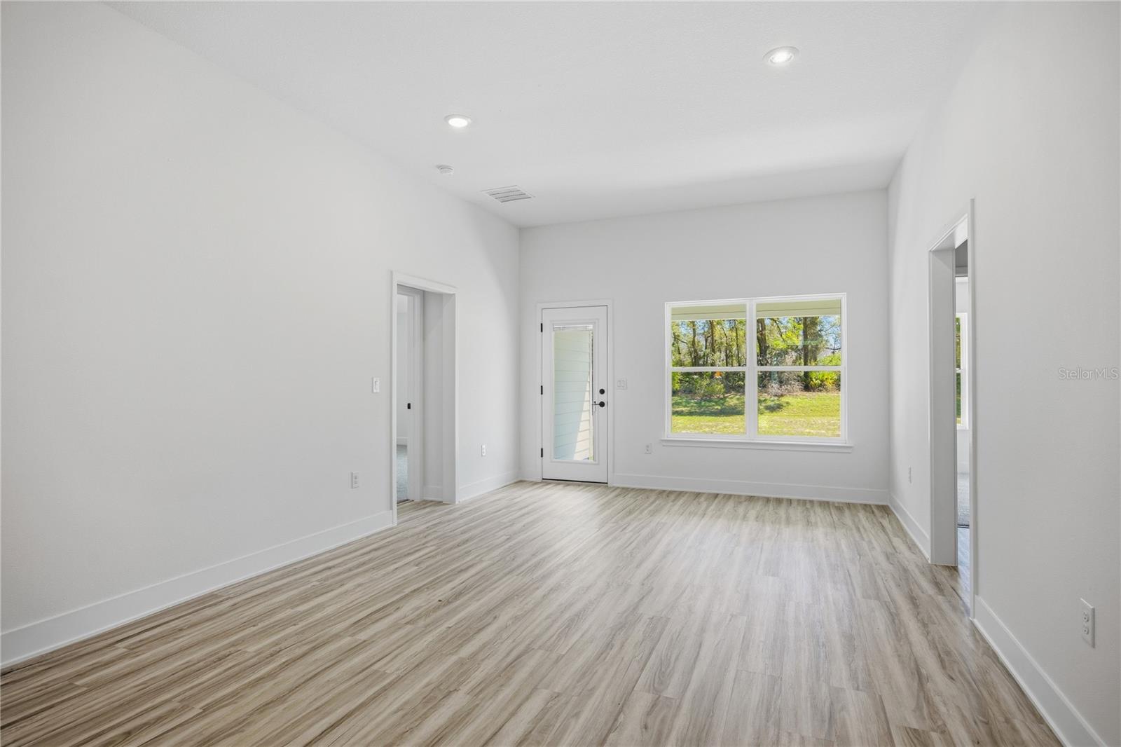 Empty, bright living room with white walls, light wood-look laminate flooring, and a glass door leading to a backyard view.