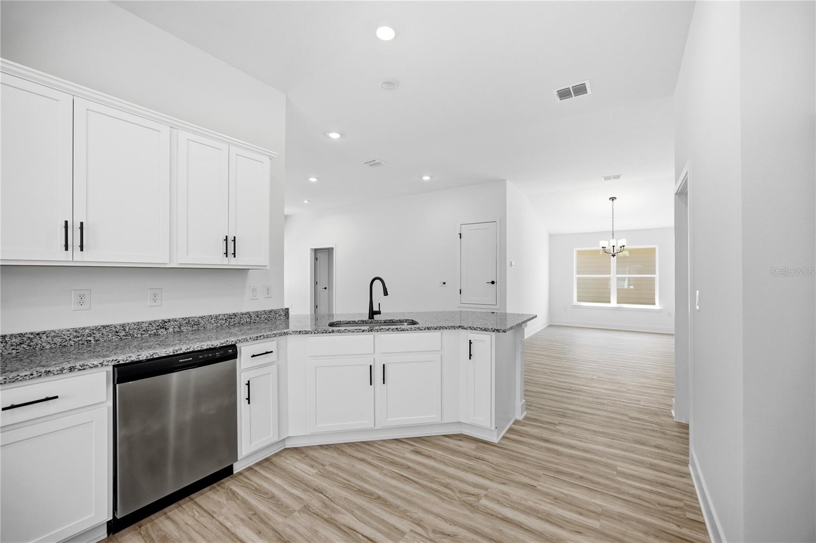 A modern kitchen with white cabinets, speckled granite countertops, and stainless steel appliances, opening into a bright dining area with wood-look flooring.