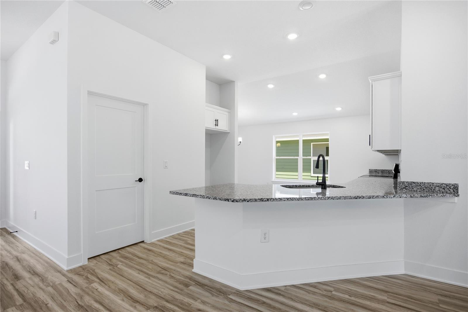 A modern, vacant kitchen with white cabinets, speckled granite countertops, and light wood-look flooring.