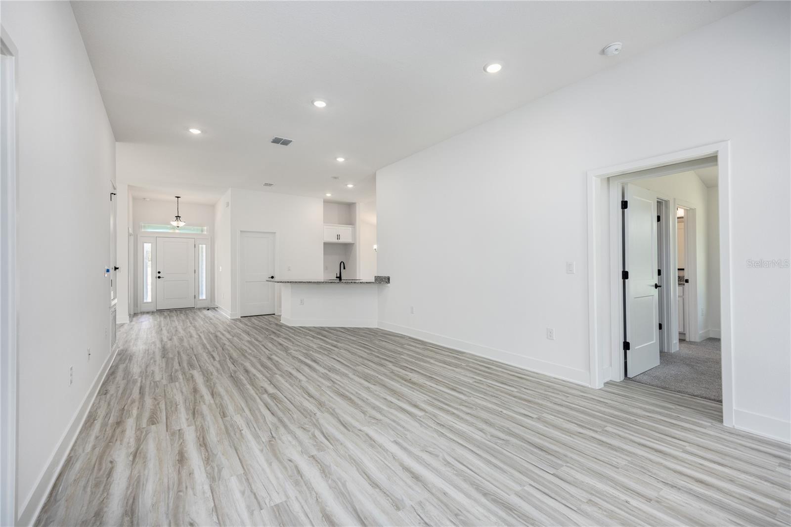 An open-plan living area featuring light wood-look floors, recessed lighting, and a view leading toward a kitchen island and front entryway.