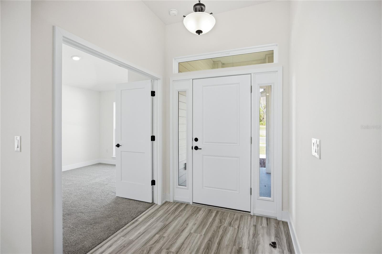 A brightly lit foyer with a white front door featuring side lites and a transom window, with a view into a carpeted room through an open doorway.