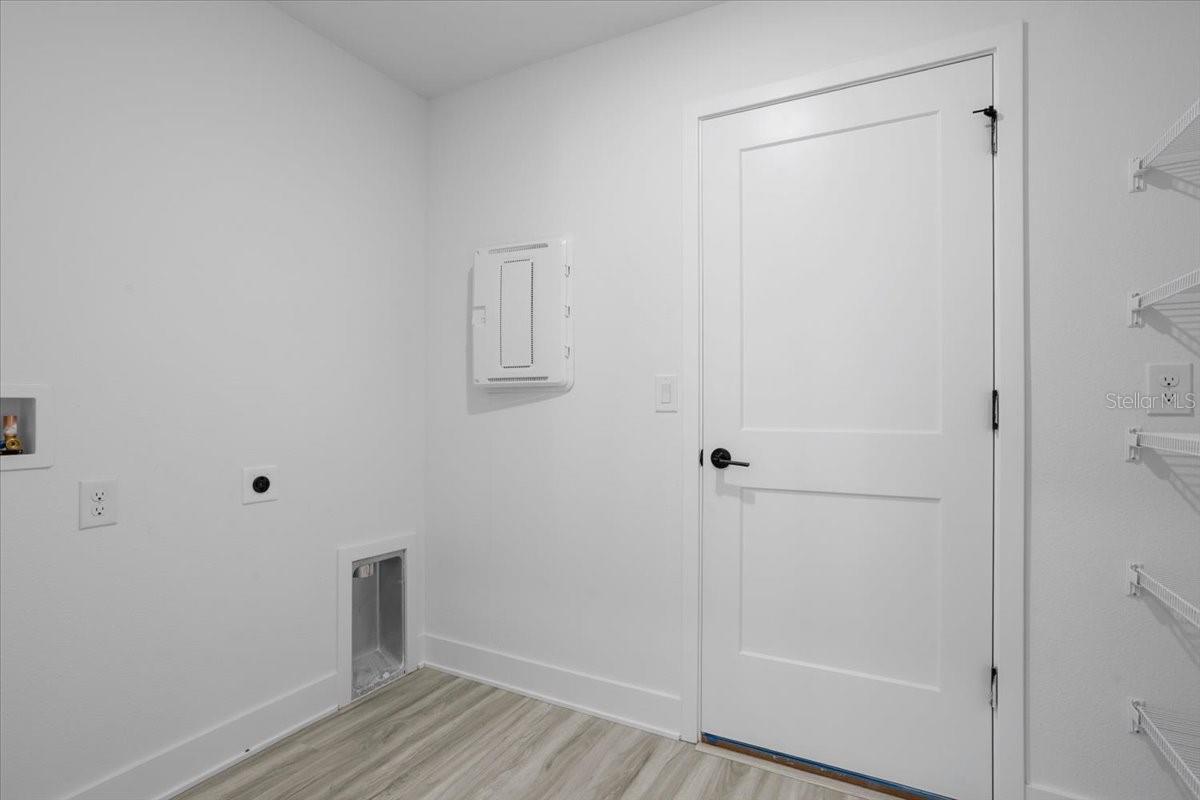 A modern laundry room with light wood floors, a white door, and utility connections on the wall.