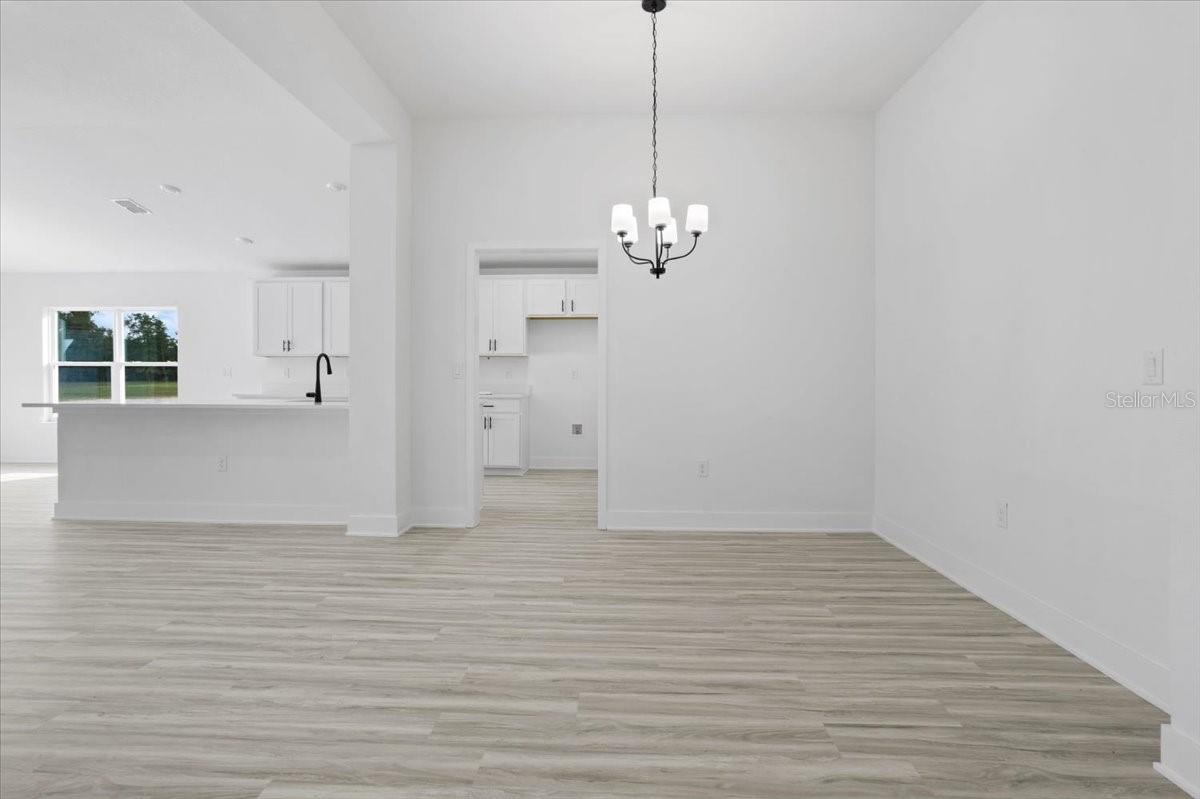 A wide angle shot of a modern, empty living room with light grey wood-look floors, white walls, and a contemporary chandelier, showing a view into the kitchen.