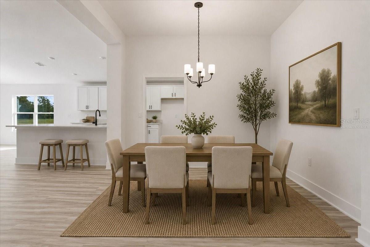 A brightly lit, modern dining room with a wooden table, six upholstered chairs, a neutral area rug, a hanging chandelier, and a view into a white kitchen.