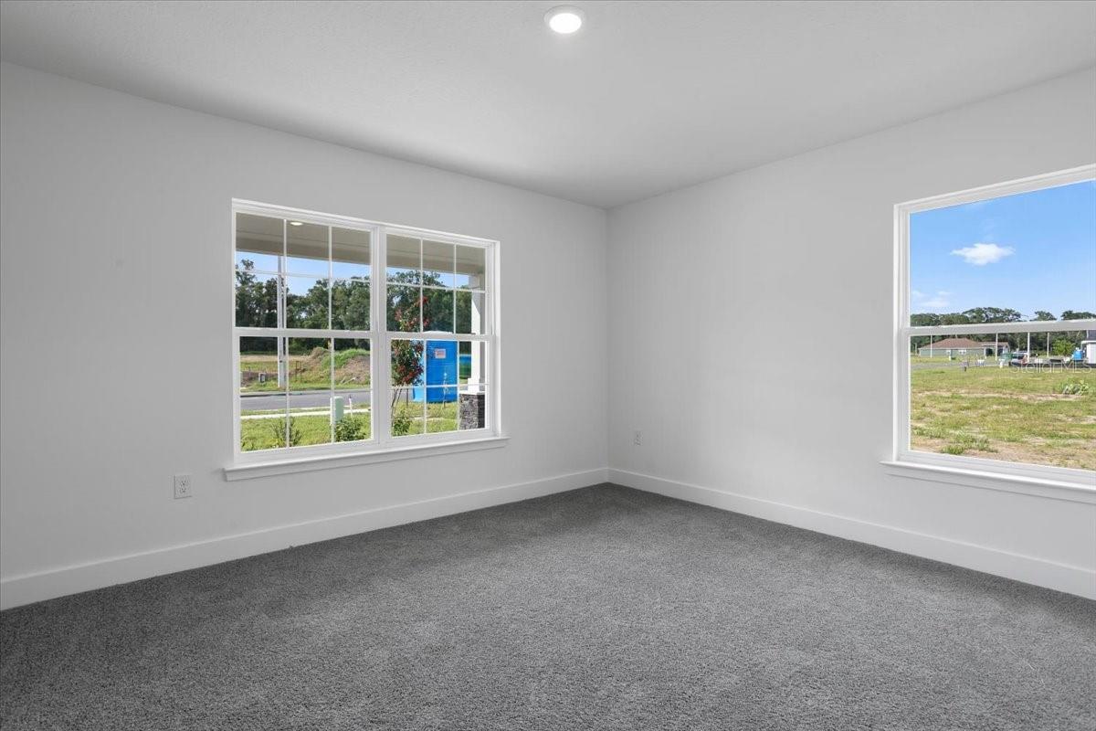 An empty bedroom with gray carpeting, white walls, and two large windows looking out onto a grassy lot.