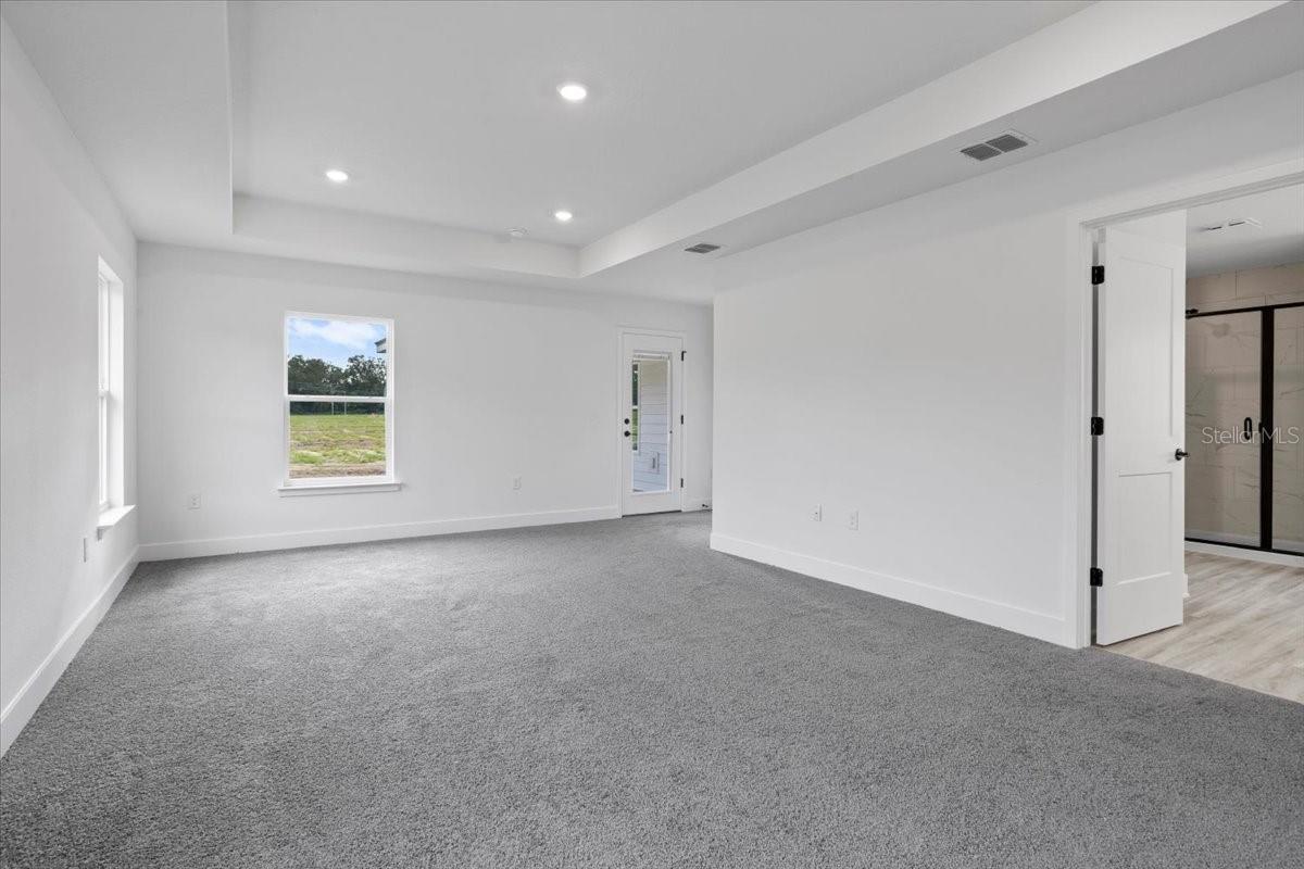 A bright, empty master bedroom featuring white walls, gray carpeting, a tray ceiling with recessed lighting, two windows, and a doorway leading into an en-suite bathroom.