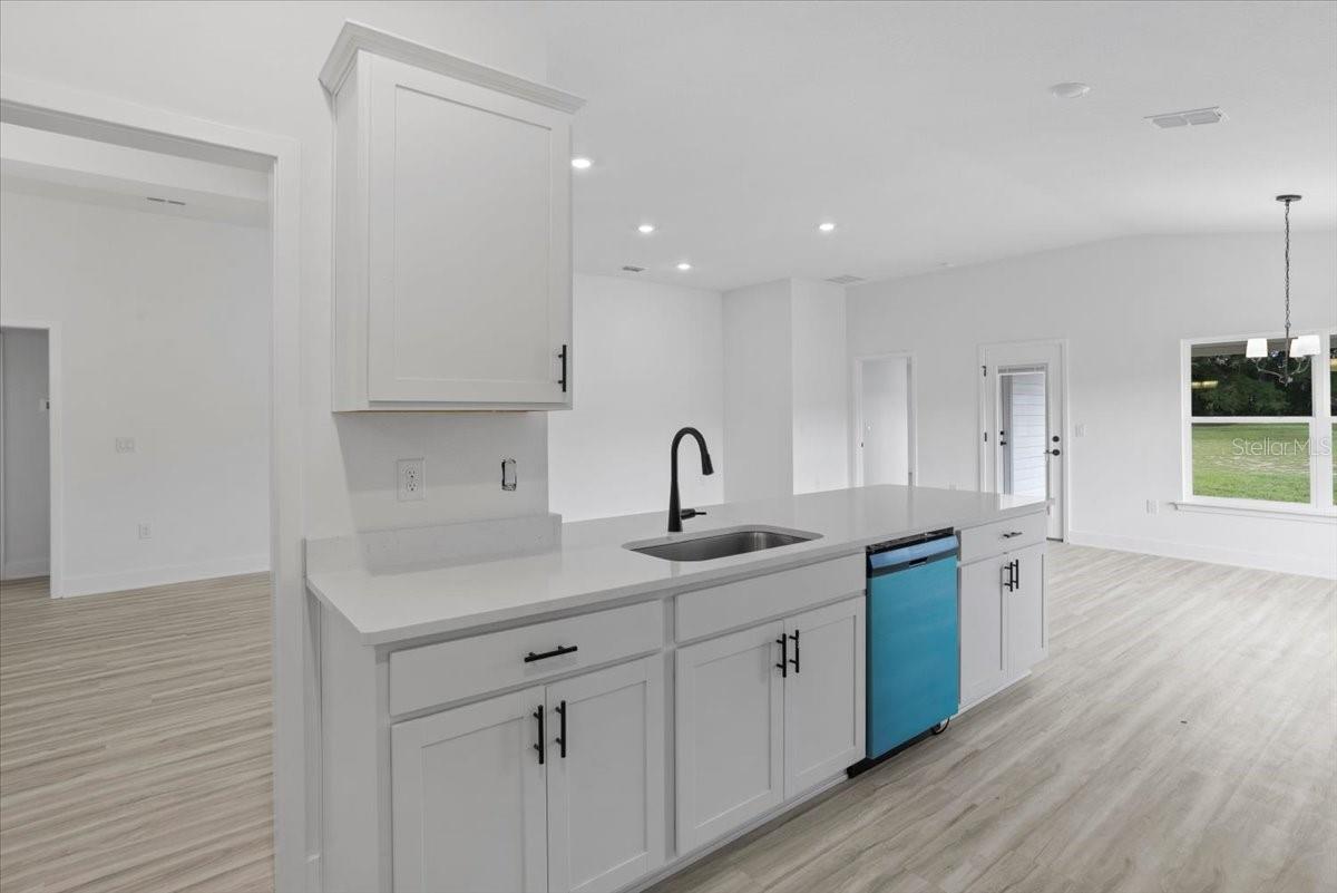A modern kitchen island with a stainless steel sink, white shaker cabinets, and a blue dishwasher in a bright open-concept home.